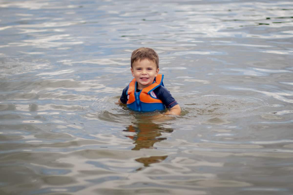Little boy in life jacket and rash guard swimming in lake 