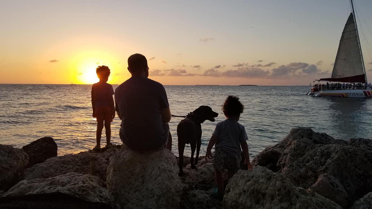 dad, two kids and dog sitting on the rocks at sunset with a boat in the distance