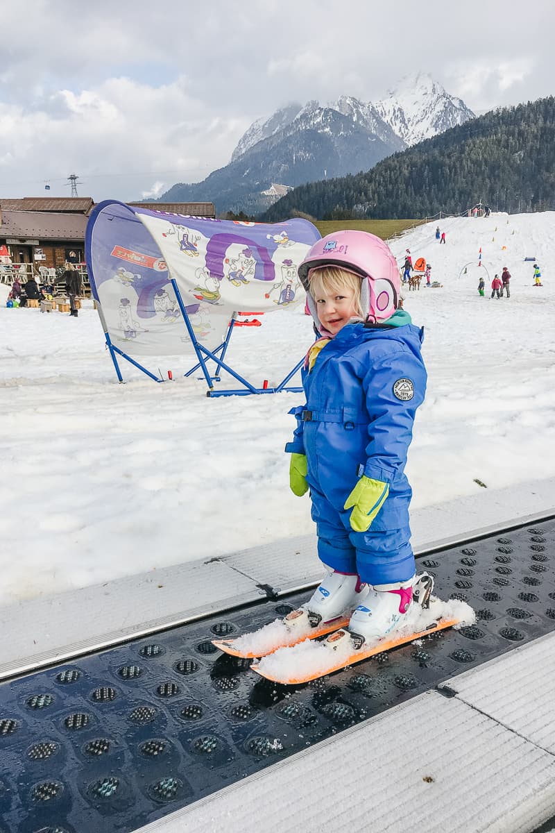 a small child takes a magic carpet up a nursery slope while she learns to downhill ski