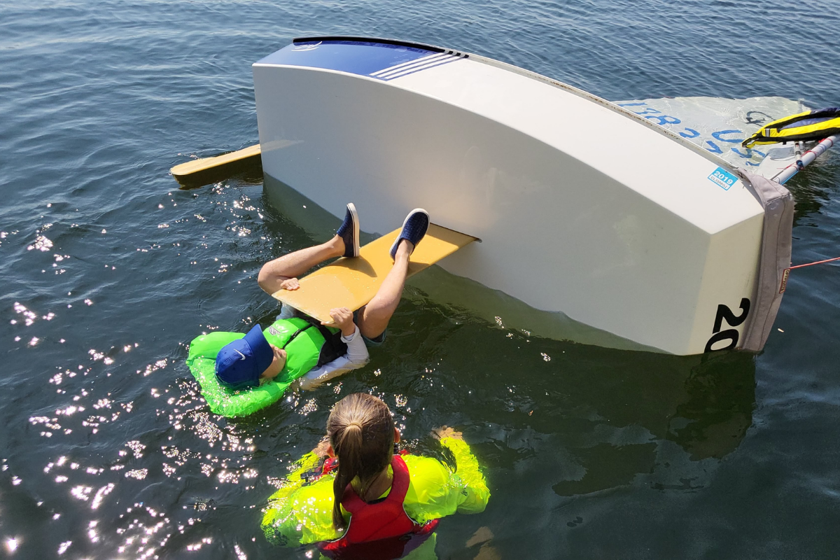A sailing instructor giving safety instruction to a kid hanging onto the keel of a capsized dinghy sailboat