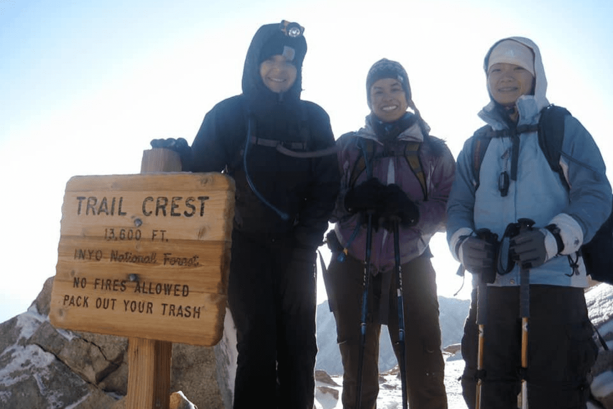Three hiking women standing at the top of a mountain in front of a sign that says 'trail crest'.