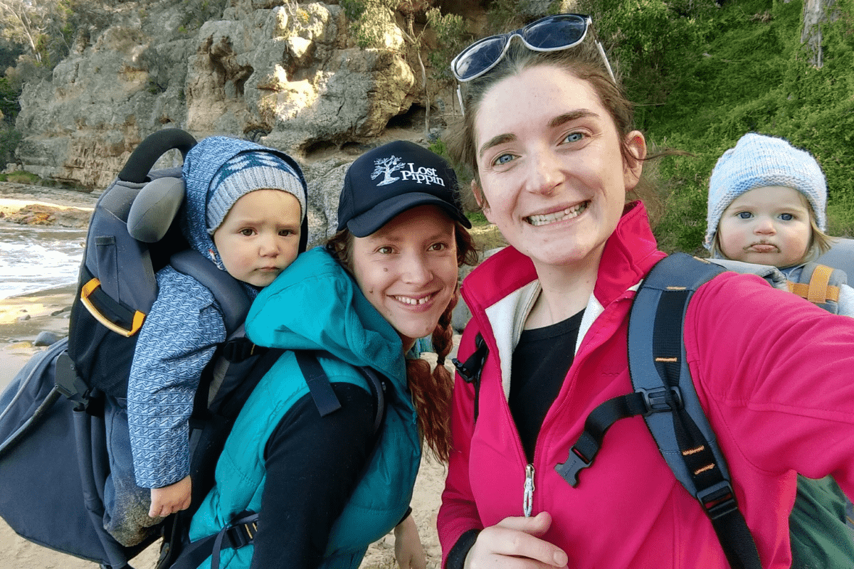 Two friends hiking at the beach with toddlers in baby carrying backpacks.