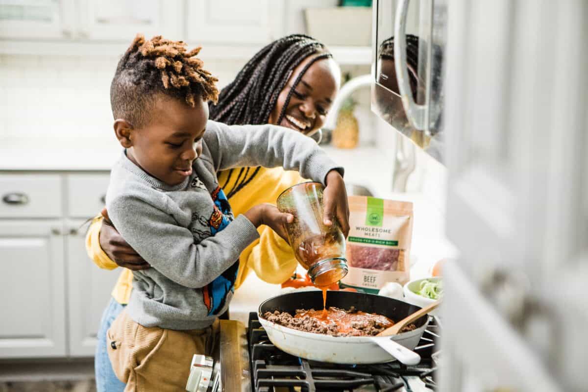 mom cooking with child and letting him pour sauce in pan - how to raise independent kids